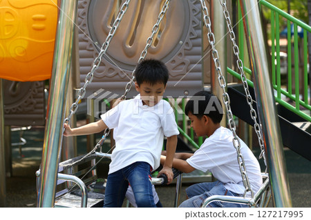 Two Young Boys Playing Together on Swings at a Colorful Playground Under Bright Sunlight in a Fun Outdoor Environment Near Vibrant Play Equipment 127217995