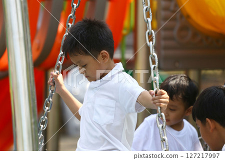 Young boy playing on swings at a playground during a sunny day, demonstrating joy and childhood innocence while engaging with friends in outdoor fun and adventure. 127217997