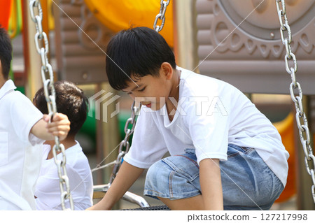 Young Boy Playing on Swing at Playground, Engaging in Fun Outdoor Activities, Joyful Child Enjoying Sunlight and Nature with Friends in Background 127217998