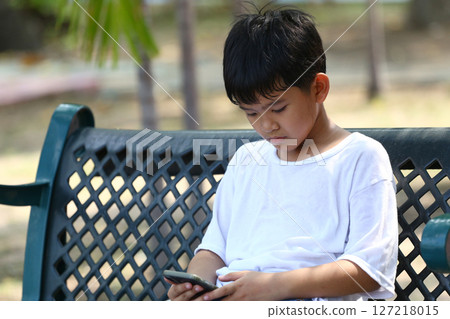 Young boy sitting alone on a park bench, focused on smartphone, showcasing the impact of technology on children's play and social interaction in everyday life. 127218015