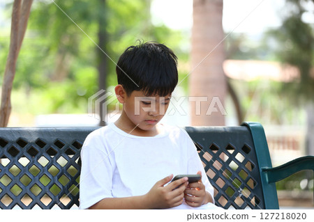 Young boy engrossed in smartphone while sitting on park bench, showcasing technology's impact on childhood and leisure activities in a natural environment Young boy engrossed in smartphone while sitting on park bench, showcasing technology's impact on childhood and leisure activities in a natural environment 127218020