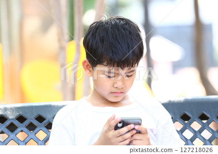 Young boy focused on smartphone in a park setting, displaying concentration and engagement while sitting on a bench with sunlight filtering through trees in the background. Young boy focused on smartphone in a park setting, displaying concentration and engagement while sitting on a bench with sunlight filtering through trees in the background. 127218026