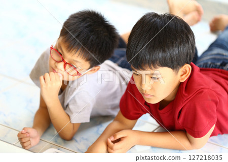 Two Young Boys Engaged in Focused Activity on Soft Floor, One Wearing Glasses and the Other with Short Hair, Bright Colors and Casual Outfits Captured in Natural Light Two Young Boys Engaged in Focused Activity on Soft Floor, One Wearing Glasses and the Other with Short Hair, Bright Colors and Casual Outfits Captured in Natural Light 127218035