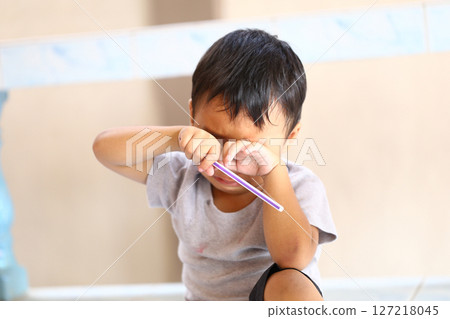 Young Child with Short Black Hair Expressing Emotion by Rubbing Eyes with Toy Brush While Sitting on Floor in Bright Indoor Environment 127218045