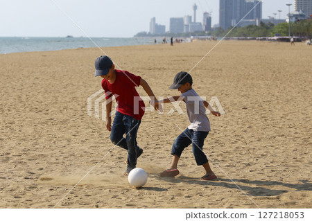 Two Young Boys Playing Soccer on Sandy Beach with Sunny Sky and Cityscape in the Background, Capturing Joy of Childhood and Outdoor Activity in a Coastal Setting Two Young Boys Playing Soccer on Sandy Beach with Sunny Sky and Cityscape in the Background, Capturing Joy of Childhood and Outdoor Activity in a Coastal Setting 127218053
