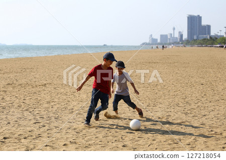 Two Young Boys Playing Soccer on a Serene Beach with City Skyline in the Background, Enjoying a Sunny Day of Fun and Friendship by the Sea Two Young Boys Playing Soccer on a Serene Beach with City Skyline in the Background, Enjoying a Sunny Day of Fun and Friendship by the Sea 127218054
