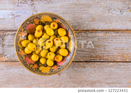 Decorative bowl filled with golden olives sits atop rustic wooden table, inviting taste of flavorful delights. Decorative bowl filled with golden olives sits atop rustic wooden table, inviting taste of flavorful delights. 127223431