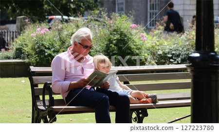 Elderly man reading to child on a sunny day in a park with blooming flowers and greenery 127223780