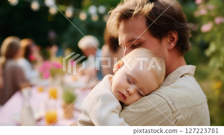 Father holding sleeping baby during outdoor gathering at sunset surrounded by friends and family 127223781