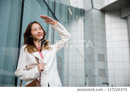Happy woman wearing white suit jacket with brown bag at sunlight outdoors. Portrait of confidence young businesswoman standing outside office building in city raise your hand to shade sun Happy woman wearing white suit jacket with brown bag at sunlight outdoors. Portrait of confidence young businesswoman standing outside office building in city raise your hand to shade sun 127226379