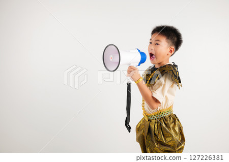 Portrait Thailand kid boy smiling traditional Thai dress costume shouting by megaphone, studio shot isolated white background, primary screeching through in megaphone announces discounts 127226381