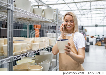 A Caucasian woman examines a flowerpot while talking on her phone inside a plant shop. Various other flowerpots are on shelves in the background. 127226910