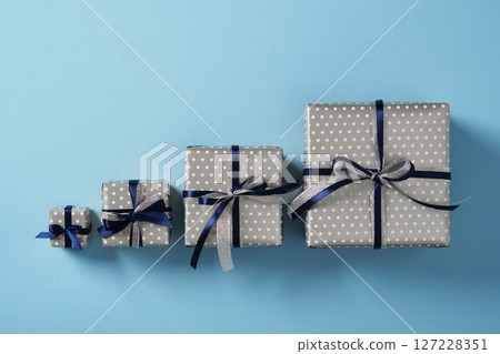 Four gift boxes of different sizes are lined up on a pastel blue background. Each gift is decorated with dark blue and silver satin ribbons. 127228351