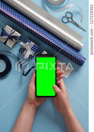 Smartphone with green screen in hands above a table with gift wrapping items. Rolls of wrapping paper, scissors, ribbon, tape. Space for your app mockup. 127228362