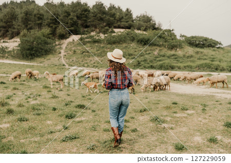 A woman wearing a hat and jeans walks through a field of sheep 127229059