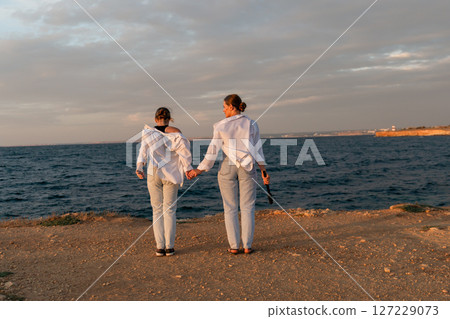 Couple Beach Sunset Holding Hands - Two women stand on a beach, holding hands and looking at the sunset. Couple Beach Sunset Holding Hands - Two women stand on a beach, holding hands and looking at the sunset. 127229073