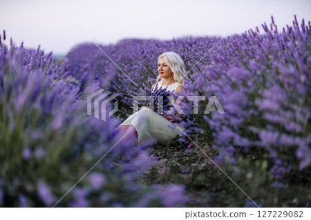 Blonde woman poses in lavender field at sunset. Happy woman in white dress holds lavender bouquet. Aromatherapy concept, lavender oil, photo session in lavender 127229082