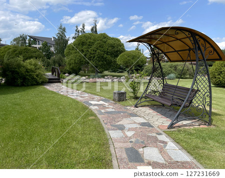 Foot path of colorful tile pieces and swings in park. Green lawn and trees in summer on sunny day. Background for design. Ecology and nature. Landscape design.  127231669
