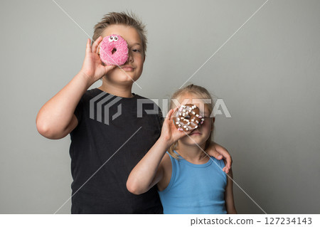 Cheerful sister and brother having fun holding donuts standing against white background wall.  127234143