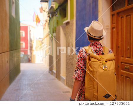 Woman in dress strolls through colorful streets of Spanish coastal town of La Vila Joiosa . sunny winter atmosphere highlights charm of Mediterranean architecture and quiet seaside life, back view 127235661