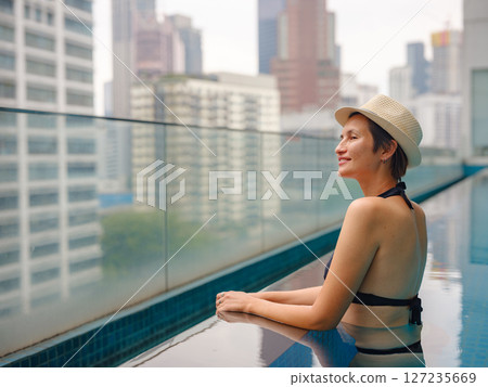 Young asian woman relaxing by pool at Kuala Lumpur hotel with view of surrounding skyscrapers, enjoying leisure time in vibrant urban setting. 127235669