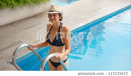Young asian woman relaxing by pool at Kuala Lumpur hotel with view of surrounding skyscrapers, enjoying leisure time in vibrant urban setting. 127235672
