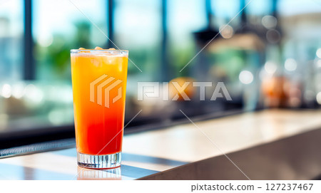 Layered orange red mocktail with ice stands alone on glossy bar counter in bright modern setting, cocktail repeated for clean refreshment, stylish bar vibe, daytime refresh concept 127237467