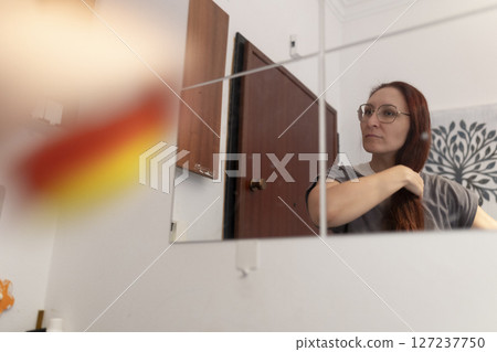 Woman styling her long hair in front of mirror at home Woman styling her long hair in front of mirror at home 127237750