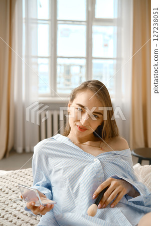 Young woman applying makeup indoors in a cozy, well-lit bedroom with natural light 127238051