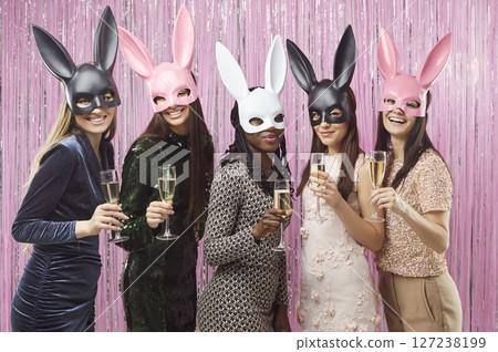Women in dress clothes and carnival bunny mask on festive pink background with champagne glasses Women in dress clothes and carnival bunny mask on festive pink background with champagne glasses 127238199