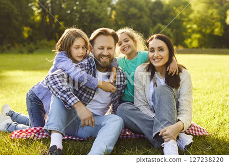 Happy family sitting together on plaid in nature, enjoying picnic time in park, smiling gladfully 127238220