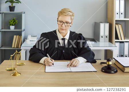 Judge serious woman in professional outfit black robe writing reviewing documents in court room 127238291