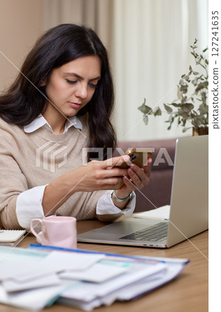 portrait of woman holds smart phone uses laptop in home office 127241635
