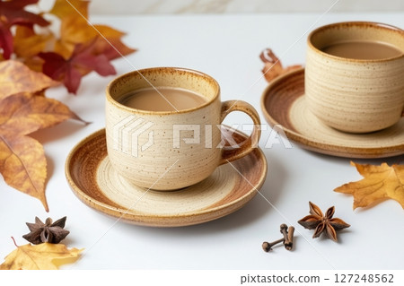 Spicy cappuccino two cups. Spice latte with art foam, cinnamon stick and anise stars. Autumn leaves. Warm Coffee on a white. Cozy fall drink on a white table. Inviting atmosphere. Mug of warming drink 127248562