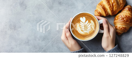 Person Hands Holding Coffee Cappuccino Cup. Composition with Coffee Latte with Milk Foam Art and Tasty Fresh Croissant on a Gray Table. Food series. Breakfast with Fresh Baked Pastry. Closeup top view 127248829