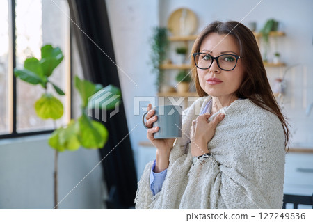 Relaxed middle aged woman with mug under blanket in home interior 127249836