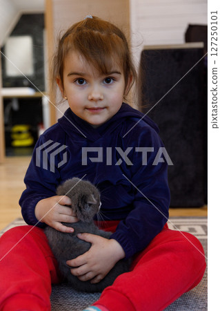 A young girl with brown hair and big eyes sits on the floor, gently holding a cute grey kitten in her arms while wearing a cozy navy sweater and vibrant red pants in a stylish room. A young girl with brown hair and big eyes sits on the floor, gently holding a cute grey kitten in her arms while wearing a cozy navy sweater and vibrant red pants in a stylish room. 127250101