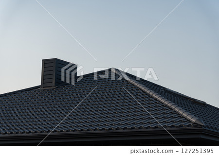 A modern house features a gray tiled roof with a prominent chimney, set against a clear blue sky during the day 127251395