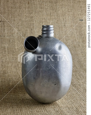 Old soldier's aluminum canteen with lid on chain on burlap. Thermos for water. Empty canteen with an unscrewed stopper. Old soldier's aluminum canteen with lid on chain on burlap. Thermos for water. Empty canteen with an unscrewed stopper. 127251491