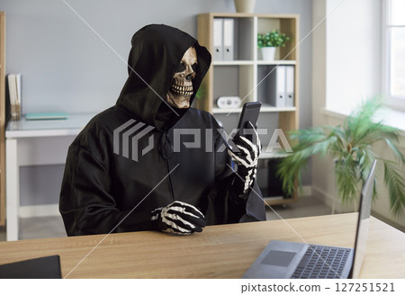 Man in death costume with black hood and skeleton mask sitting in office looking at mobile phone 127251521