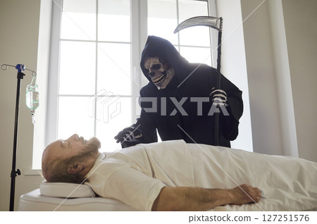 Death holding scythe standing near dying elderly senior man lying in medical clinic under IV drip. 127251576
