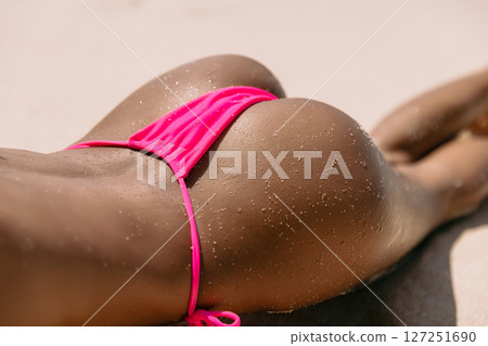 Close up of slim woman in a pink bikini lying on the sandy beach with sun-kissed skin and water droplets. Close up of slim woman in a pink bikini lying on the sandy beach with sun-kissed skin and water droplets. 127251690