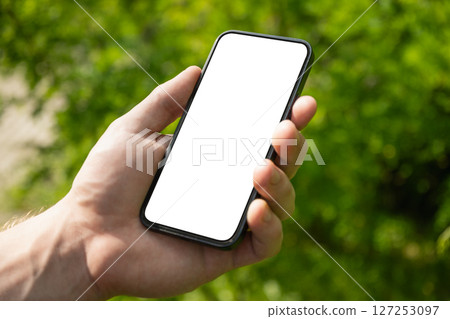 Man hand holding mobile phone with white blank screen against blurred green background. Showcasing mobile apps, design mockups, or digital content presentations. Check apps. Summer heat weather. Man hand holding mobile phone with white blank screen against blurred green background. Showcasing mobile apps, design mockups, or digital content presentations. Check apps. Summer heat weather. 127253097