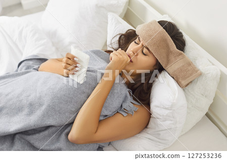 Sick young woman with wet towel on head lying in bed, suffering from cold or flu virus and coughing. 127253236
