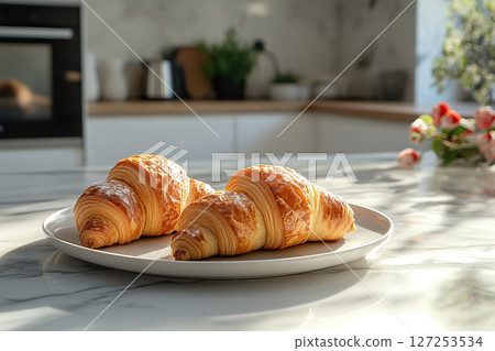 Two delicious french croissants on a plate on a table in a modern kitchen in the rays of the morning sun. 127253534