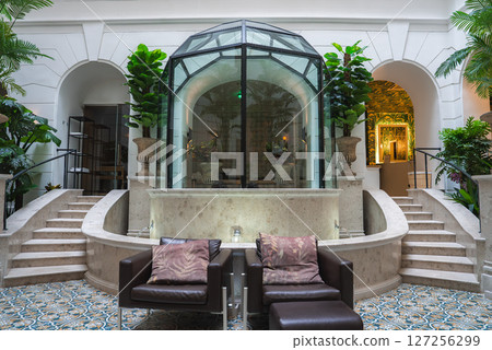 Luxury hotel interior featuring a glass atrium, lush greenery in ornate planters, symmetrical staircases, and a cozy seating area with leather armchairs. Luxury hotel interior featuring a glass atrium, lush greenery in ornate planters, symmetrical staircases, and a cozy seating area with leather armchairs. 127256299