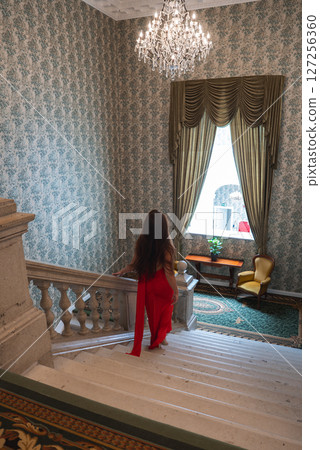 A beautiful woman in a red dress descends a marble staircase with a classic balustrade. Floral wallpaper, a chandelier, and yellow armchairs enhance the decor. 127256360