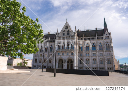 The Gothic Revival style Hungarian Parliament Building in Budapest, featuring spires and ornate windows, with a tree and paved area in the foreground. The Gothic Revival style Hungarian Parliament Building in Budapest, featuring spires and ornate windows, with a tree and paved area in the foreground. 127256374