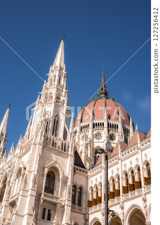 The Hungarian Parliament Building in Budapest displays intricate spires, a large central dome, and ornate detailing under a clear blue sky. 127256412