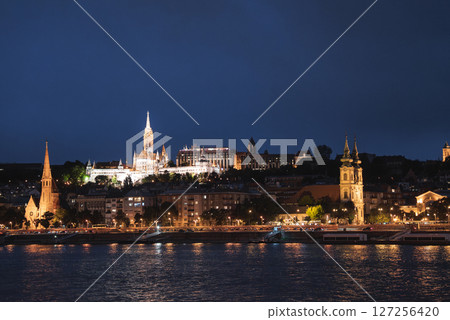 Illuminated Matthias Church and Fisherman's Bastion atop Castle Hill in Budapest, Hungary, with the Danube River reflecting city lights at night. Illuminated Matthias Church and Fisherman's Bastion atop Castle Hill in Budapest, Hungary, with the Danube River reflecting city lights at night. 127256420